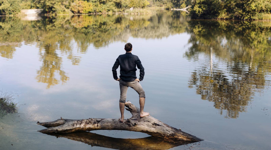 Christian Ploy steht selbstbewusst auf einem Baumstamm in der Mitte eines Gewässers mit viel Natur